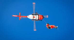 Blick von unten auf einen fliegenden rot-weißen Rettungshubschrauber mit einer Person an der Seilwinde hängend