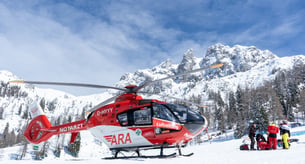 Ein rot-weißer Hubschrauber steht auf einer Skipiste, daneben wird eine verunfallte Person versorgt