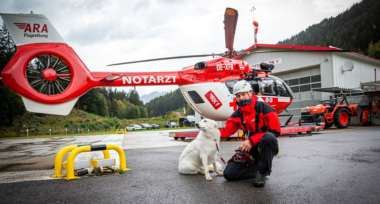 Ein Bergretter kniet neben einem Hund mit weißem Fell vor einem rot-weißen Rettungshubschrauber