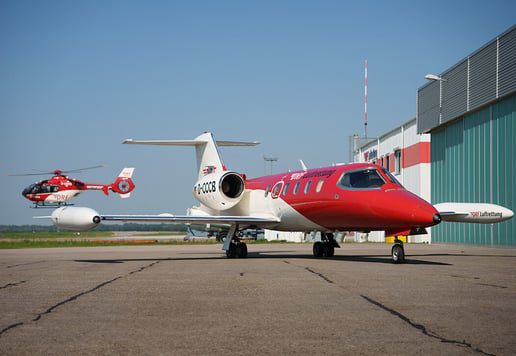 Ein rot-weißer Learjet der DRF Luftrettung steht auf einem Flugplatz, im Hintergrund hebt ein Rettungshubschrauber ab.